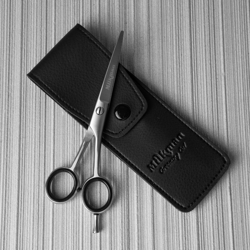 A pair of silver beard and mustache trimming scissors with a dark silver finish, placed next to a black vegan leather pouch with the Milkman logo.